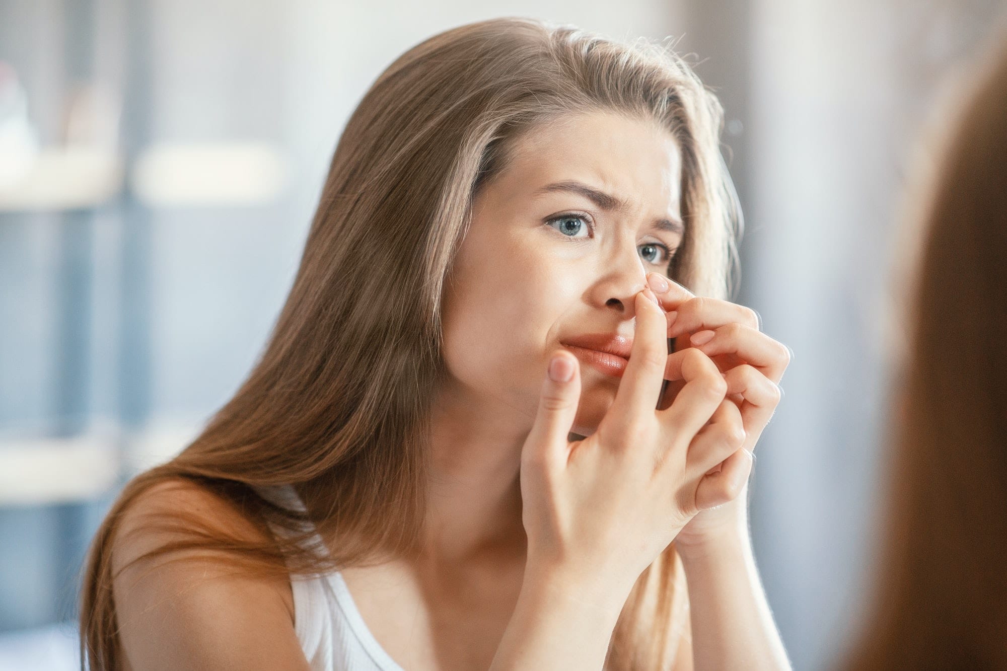 Displeased young woman squeezing acne on her nose while looking in mirror indoors