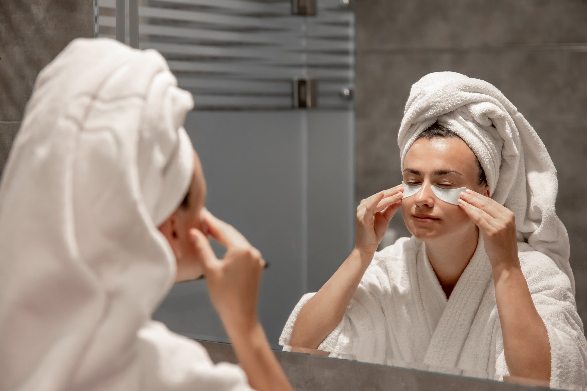 A young woman in a robe and with a towel on her head is putting patches under her eyes.