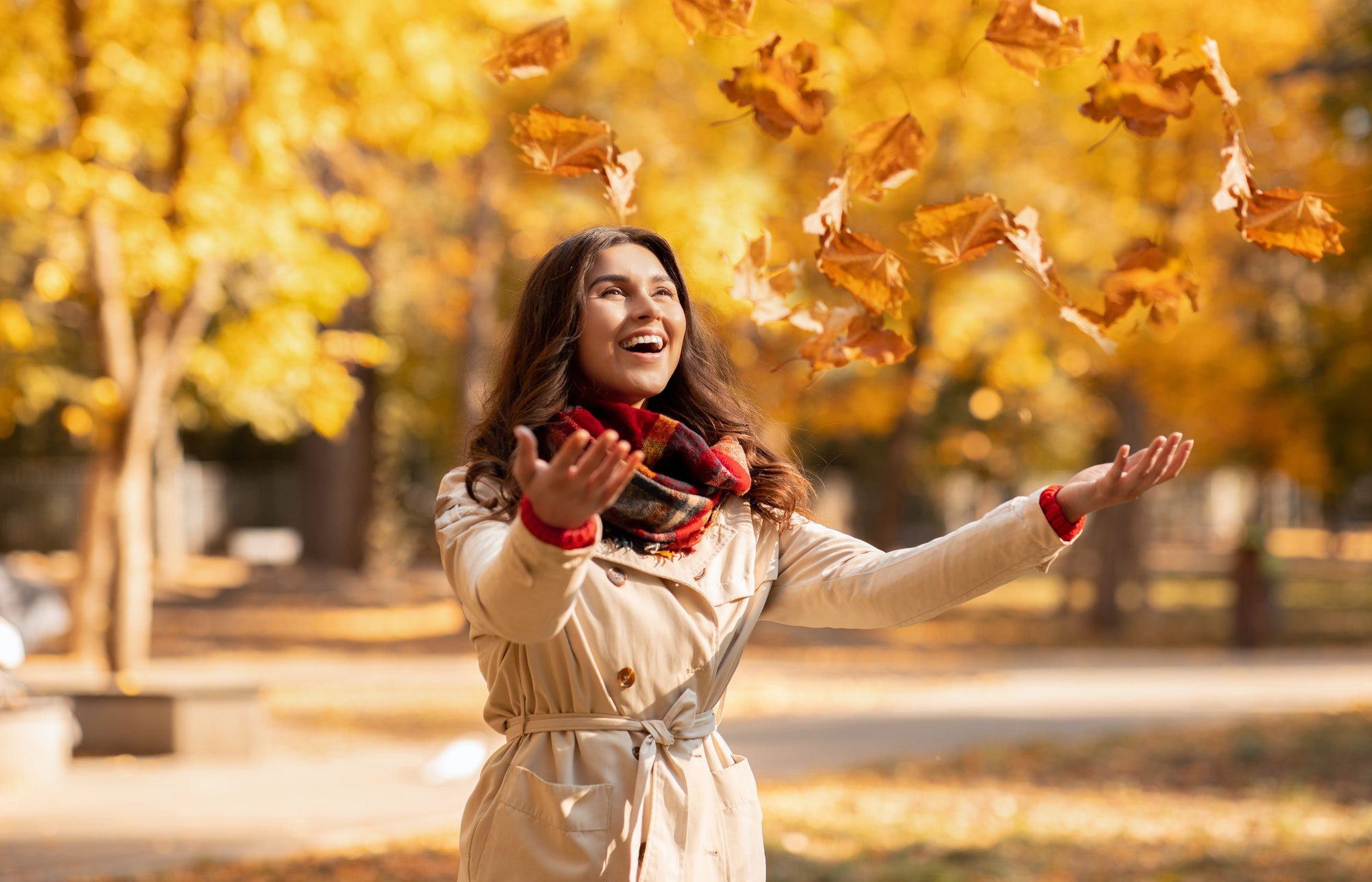 Joyful young woman in autumn outfit catching yellow leaves during her walk at park on bright fall