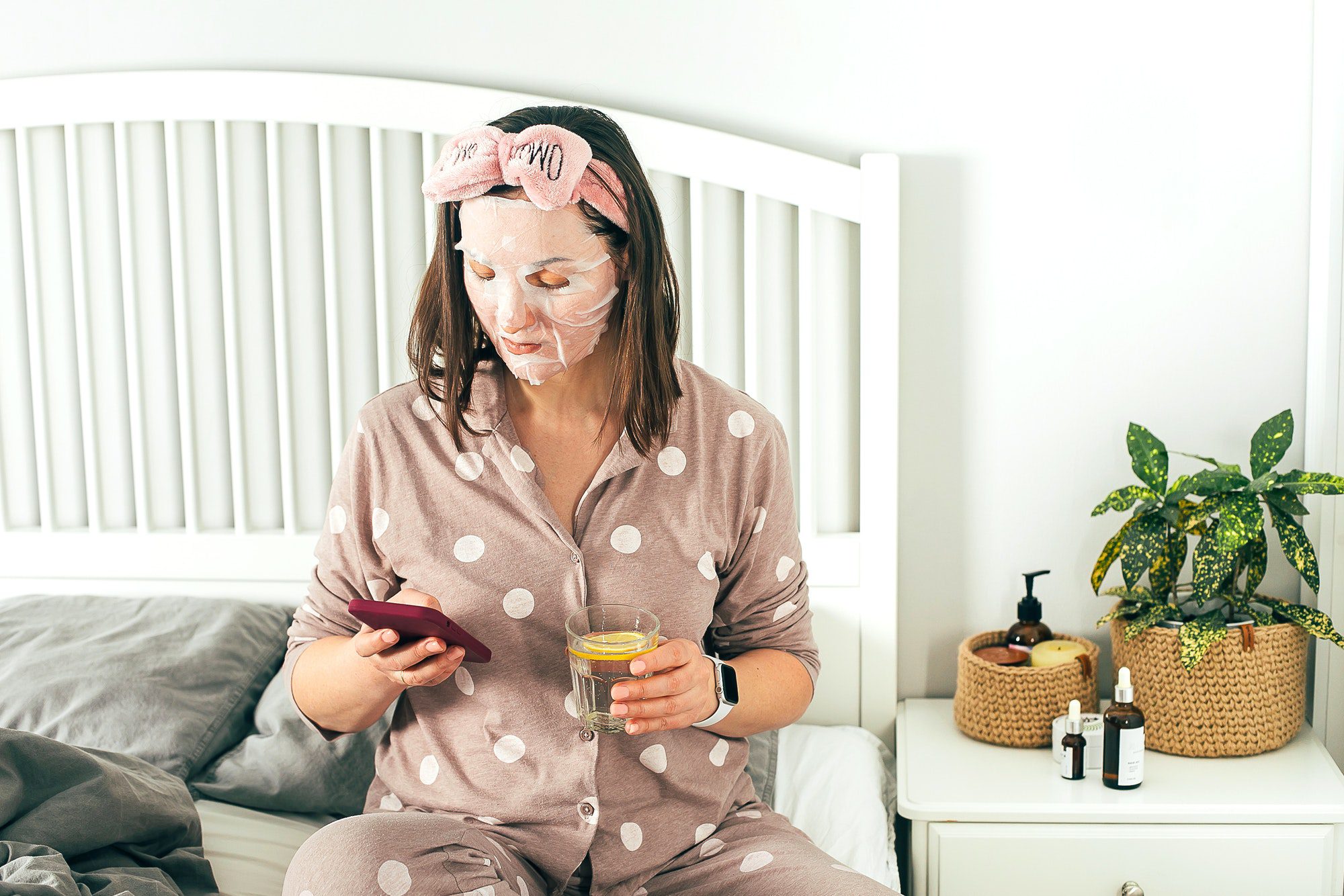 Young woman with sheet mask. Adult woman using phone, drink water. Morning routine, selfcare concept