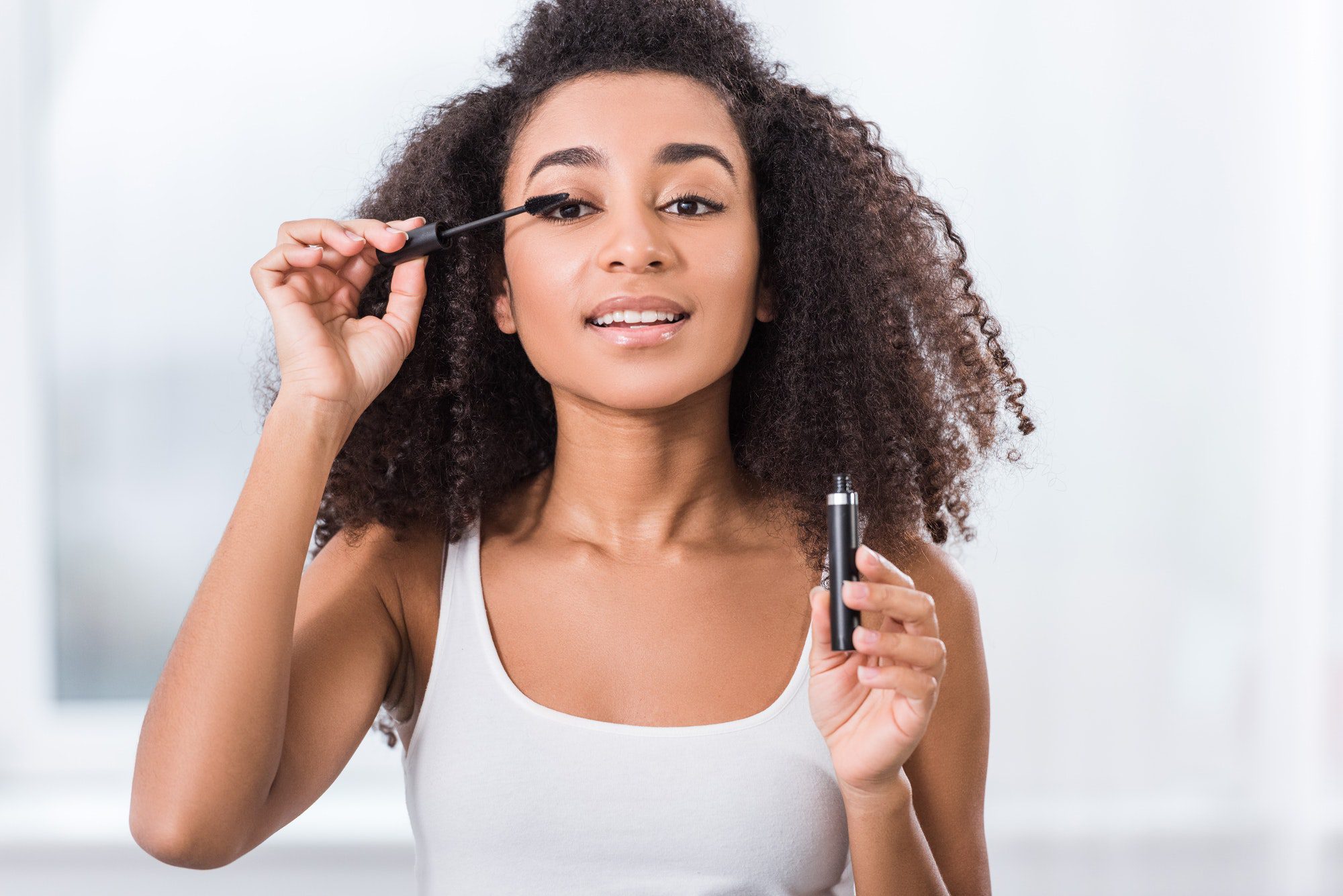 curly african american girl using mascara and looking at camera