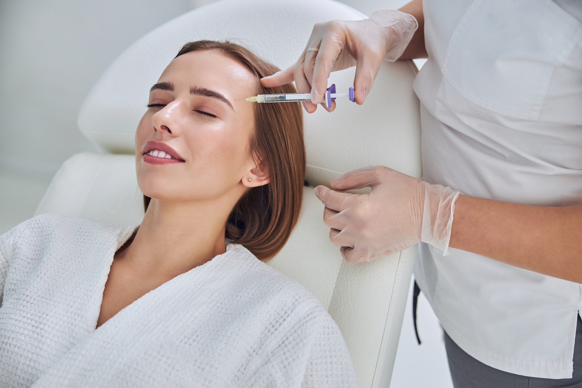 Adorable pretty lady in white bathrobe receiving plastic surgery injection on her face in cosmetic