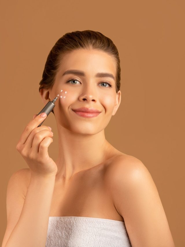 Beautiful young woman with silky skin applying face cream or cosmetic highlighter on brown studio