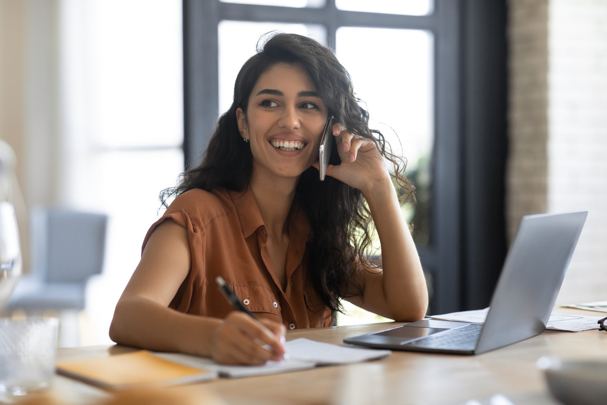 Cheerful young businesswoman talking on cellphone, using laptop, working remotely from home. Modern