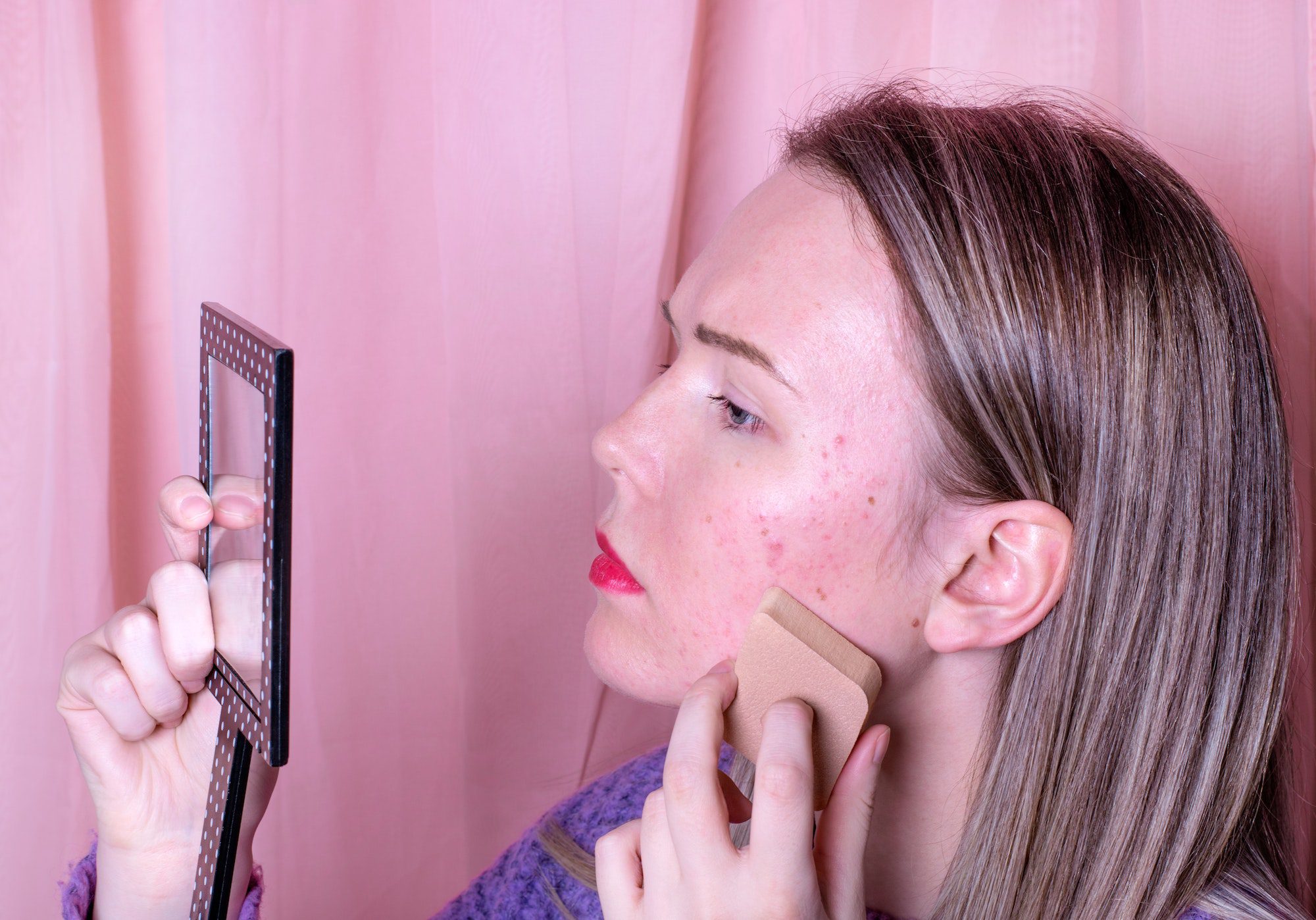 Young woman is covering acne by applying liquid foundation with makeup sponge looking into mirror